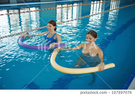 Two young women training with noodles at swimming pool Two young women training with noodles at swimming pool 107897076