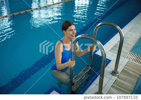 Happy smiling woman climbing out by steps of swimming pool Happy smiling woman climbing out by steps of swimming pool 107897110