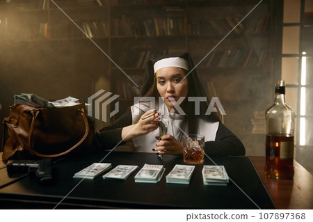 Pretty confident woman in nun dress sitting at table with money stacks 107897368