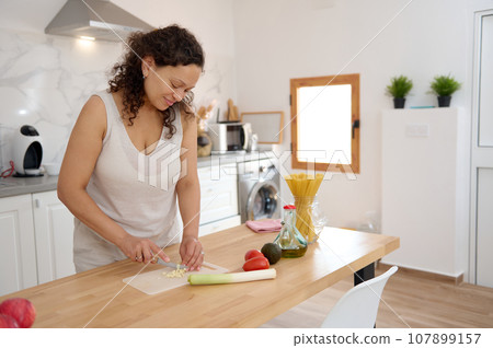 Young adult Latin American woman cutting garlic, preparing a sauce for Italian pasta in the home kitchen 107899157