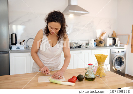 Latin American female chef preparing a healthy salad for dinner at home, chopping garlic on a cutting board 107899158