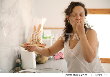 Latin American curly woman kisses fingers, shows tasty hands sign, bellissimo while cooking gourmet food in the home k 107899166