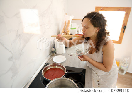 Young Latin American pretty housewife tasting the tomato sauce she's cooking in the home kitchen 107899168