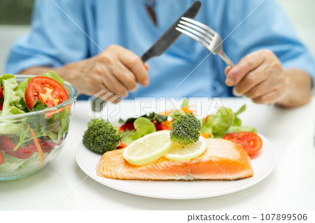 Asian elderly woman patient eating Salmon steak breakfast with vegetable healthy food while sitting and hungry on bed in hospital. Asian elderly woman patient eating Salmon steak breakfast with vegetable healthy food while sitting and hungry on bed in hospital. 107899506