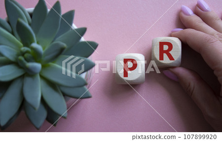 PR - Public Relations symbol. Concept word PR on wooden cubes. Businessman hand. Beautiful pink background with succulent plant. Business and PR concept. Copy space. 107899920
