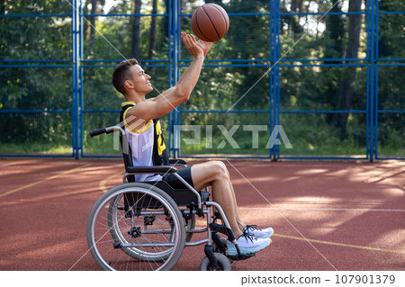 Happy basketball player with disability uses wheelchair while playing on outdoor sports court. 107901379