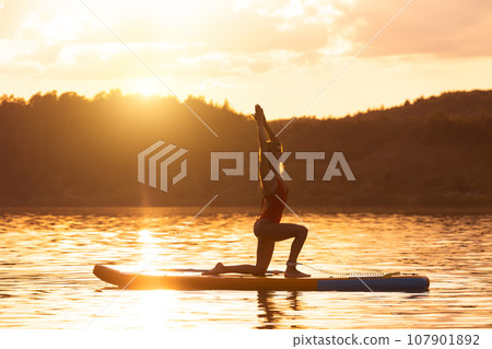 Young woman in yoga pose practicing on paddle sup surfboard during sunrise or sunset. Young woman in yoga pose practicing on paddle sup surfboard during sunrise or sunset. 107901892