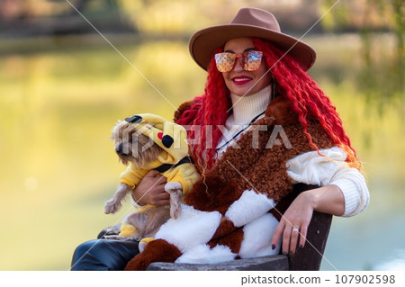 Autumn lake woman red hair dog. She sits by a pond on a wooden pier in autumn and admires nature, holding a Yorkshire terrier in her hands. The concept of tourism, weekends outside the city. Autumn lake woman red hair dog. She sits by a pond on a wooden pier in autumn and admires nature, holding a Yorkshire terrier in her hands. The concept of tourism, weekends outside the city. 107902598