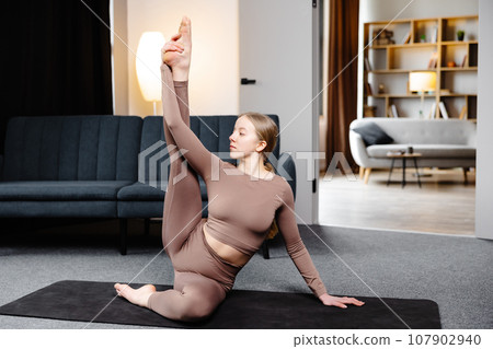 Young sporty woman practicing yoga, doing Compass pose, wearing sportswear pants and top sitting on the floor in living room Young sporty woman practicing yoga, doing Compass pose, wearing sportswear pants and top sitting on the floor in living room 107902940