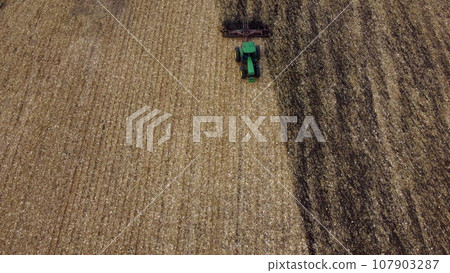 Tractor plowing the ground. Flying over green tractor that plows up ground in yellow field after harvesting wheat on autumn day. Tractor digging land. Agricultural work on field. Aerial drone view Tractor plowing the ground. Flying over green tractor that plows up ground in yellow field after harvesting wheat on autumn day. Tractor digging land. Agricultural work on field. Aerial drone view 107903287
