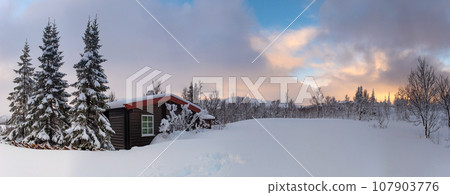 A small snow covered hunting lodge in the norwegian mountains with animal traces in the foreground and mountain summits in the background 107903776