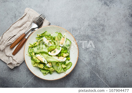 Healthy green avocado salad bowl with boiled eggs, sliced cucumbers, edamame beans, olive oil and herbs on ceramic plate top view, grey stone rustic table background. Copy space 107904477