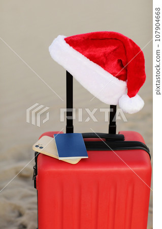 Santa Claus hat, smartphone and passport on red suitcase on seashore sand beach. Travel on Christmas and New Year winter holidays concept. Last minute trips, hot tours. Copy space for advertising 107904668