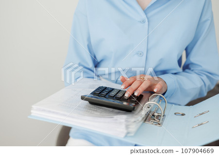 Cropped photo of female accountant in blue shirt calculates on calculator costs, counts money, company budget, analyses financial reports, checks bills and taxes. Investment, savings, currency 107904669