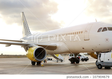 Side view of large white passenger airplane parked at terminal airport on runway against dramatic sunset background, getting ready to fly. Concept of business, economy and first class traveling Side view of large white passenger airplane parked at terminal airport on runway against dramatic sunset background, getting ready to fly. Concept of business, economy and first class traveling 107904679