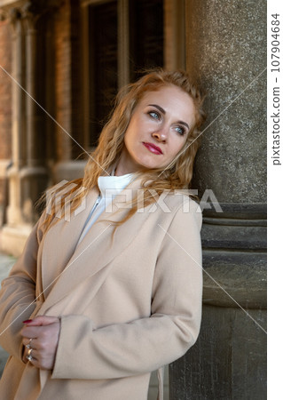 Stylish elegant young woman in beige trench coat with long red hair outside near gray column looking into the distance. Vertical frame. 107904684