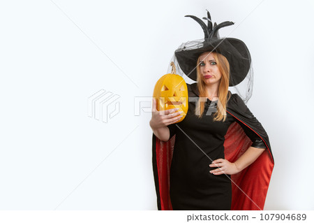 Dissatisfied young woman in witch hat with jack-o-lantern in her hands. Black clothes. Halloween party. White background, space for text Dissatisfied young woman in witch hat with jack-o-lantern in her hands. Black clothes. Halloween party. White background, space for text 107904689