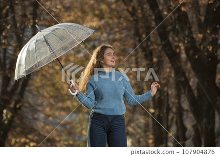 Pretty girl with long hair walks through autumn park. Young woman in blue sweater with transparent umbrella in hands Pretty girl with long hair walks through autumn park. Young woman in blue sweater with transparent umbrella in hands 107904876