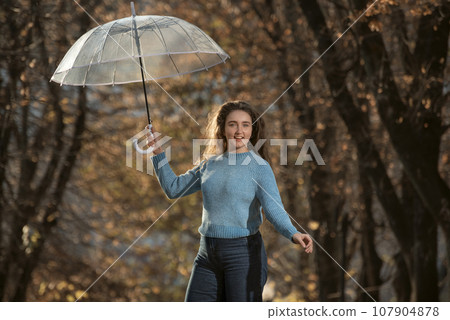 Dancing pretty girl with long hair walks through autumn park. Young woman in blue sweater with transparent umbrella Dancing pretty girl with long hair walks through autumn park. Young woman in blue sweater with transparent umbrella 107904878