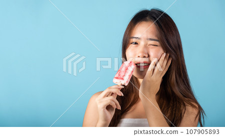 Portrait of Asian young woman with sensitive teeth after eating delicious ice cream wood stick mixed fruit flavor feeling painful uncomfortable, studio shot isolated on blue background, dental problem 107905893