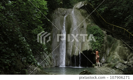 Woman near waterfall located in summer green forest. Creative. Woman walking barefoot towards the waterfall near the picturesque touristic wooden stairs. Woman near waterfall located in summer green forest. Creative. Woman walking barefoot towards the waterfall near the picturesque touristic wooden stairs. 107906271