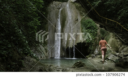Woman near waterfall located in summer green forest. Creative. Woman walking barefoot towards the waterfall near the picturesque touristic wooden stairs. Woman near waterfall located in summer green forest. Creative. Woman walking barefoot towards the waterfall near the picturesque touristic wooden stairs. 107906274