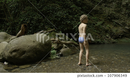 Boy walking in the forest with naked feets in the water. Creative. Boy in summer stream enjoying beautiful landscape. 107906349