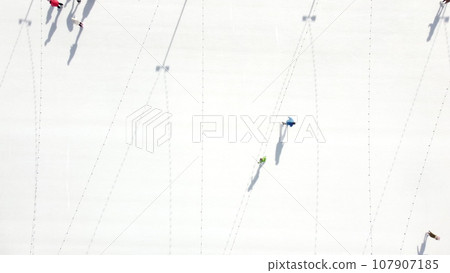People ride on white ice rink on street and cast long shadows. People skating on sunny winter day. Top view. Aerial drone view. Leisure entertainment vacation weekend holidays activities lifestyle. 107907185