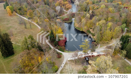 Beautiful scenery view of an autumn park with trees with yellow fallen leaves, lakes,waterfall, architecture, glades and people walking dirt paths on autumn day. Flying over the autumn park. Top view. Beautiful scenery view of an autumn park with trees with yellow fallen leaves, lakes,waterfall, architecture, glades and people walking dirt paths on autumn day. Flying over the autumn park. Top view. 107907201