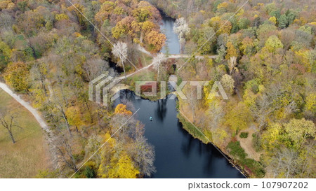 Beautiful scenery view of an autumn park with trees with yellow fallen leaves, lakes, architecture, glades and people walking along dirt paths on an autumn day. Flying over the autumn park. Top view. Beautiful scenery view of an autumn park with trees with yellow fallen leaves, lakes, architecture, glades and people walking along dirt paths on an autumn day. Flying over the autumn park. Top view. 107907202