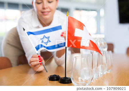 Young woman puts flags of Israel and Switzerland on table in office 107909021