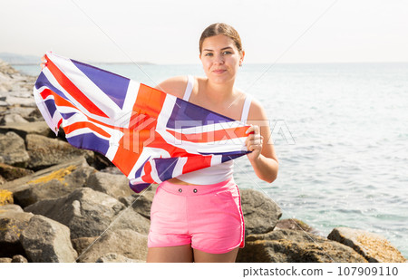 Attractive smiling woman holding Great Britain flag on the seashore on sunny day 107909110