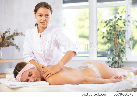 Young professional masseuse performing back massage to female client Young professional masseuse performing back massage to female client 107909173