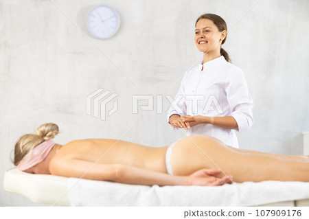 Smiling young female masseur in white coat standing in massage room Smiling young female masseur in white coat standing in massage room 107909176