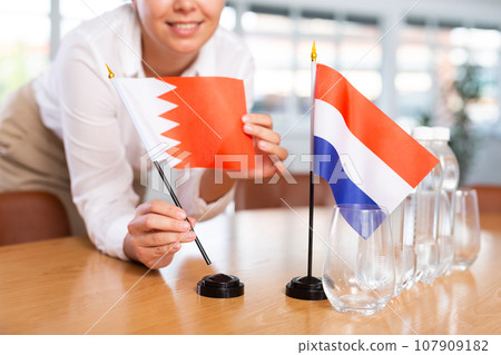 Woman secretary prepares an office for negotiations - she places flags of Netherlands and Bahrain table 107909182