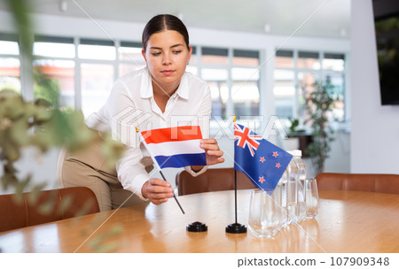 Woman secretary prepares an office for negotiations - she places flags of Netherlands and Australia table 107909348