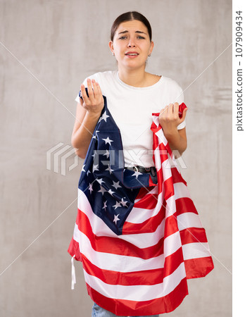 Sad young woman holding USA flag against unicoloured background 107909434