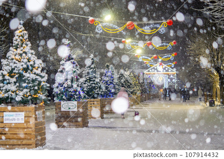 City street during snowfall at winter night. Beautiful illumination and decoration on street. New Year Christmas holidays celebration. Lanterns garlands on trees. 107914432
