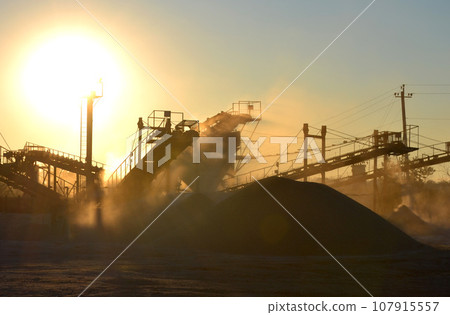 Mining quarry. Crushing plant in granite quarry at sunset dawn. Crusher in quarry. Lot of dust. Stone crushing. Industrial production of gravel. Gravel conveyor. Cone type rock crusher, shining sun Mining quarry. Crushing plant in granite quarry at sunset dawn. Crusher in quarry. Lot of dust. Stone crushing. Industrial production of gravel. Gravel conveyor. Cone type rock crusher, shining sun 107915557