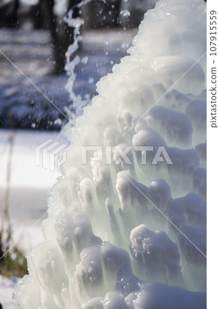 Iced fountain in the park on a winter sunny day. Fountain with frozen water. A jet of water from a fountain that iced over in the cold. Background covered with ice and snow in the park on day Iced fountain in the park on a winter sunny day. Fountain with frozen water. A jet of water from a fountain that iced over in the cold. Background covered with ice and snow in the park on day 107915559