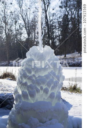 Iced fountain in the park on a winter sunny day. Fountain with frozen water. A jet of water from a fountain that iced over in the cold. Background covered with ice and snow in the park on day Iced fountain in the park on a winter sunny day. Fountain with frozen water. A jet of water from a fountain that iced over in the cold. Background covered with ice and snow in the park on day 107915569