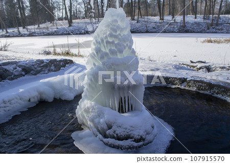 Iced fountain in the park on a winter sunny day. Fountain with frozen water. A jet of water from a fountain that iced over in the cold. Background covered with ice and snow in the park on day Iced fountain in the park on a winter sunny day. Fountain with frozen water. A jet of water from a fountain that iced over in the cold. Background covered with ice and snow in the park on day 107915570