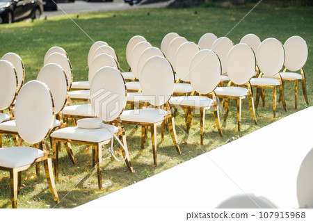Luxury white-golden chairs on wedding ceremony outdoors. Festive decorations. Empty rows armchairs for guests. Luxury white-golden chairs on wedding ceremony outdoors. Festive decorations. Empty rows armchairs for guests. 107915968