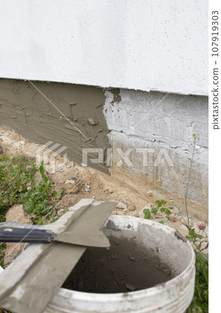 Vertical photo of a bucket with cement and a spatula against the background of the foundation of a house. 107919303