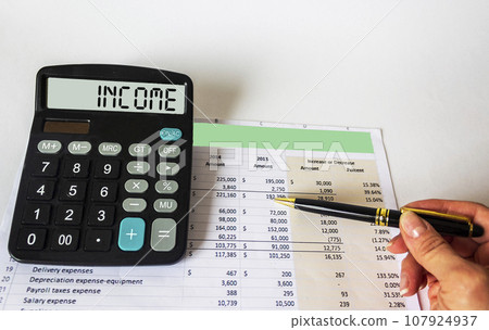 Calculator with the text INCOME on the background of documents. The female hand shows  107924937