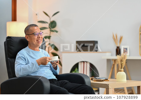 Pleased elderly man relaxing on armchair and drinking tea enjoying stress free peaceful mood wellbeing in living room. Pleased elderly man relaxing on armchair and drinking tea enjoying stress free peaceful mood wellbeing in living room. 107928899