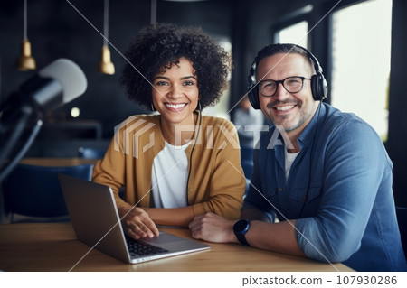Cheerful smiling multiracial blogger couple hosting a podcast in the studio. An African American woman and a Caucasian man wearing headphones sit at a table with microphones and a laptop chatting. 107930286