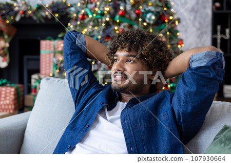 Young Indian man resting after work and study at home on sofa near Christmas tree on winter holidays. Sitting smiling and relaxed on the couch. Close-up photo. Young Indian man resting after work and study at home on sofa near Christmas tree on winter holidays. Sitting smiling and relaxed on the couch. Close-up photo. 107930664