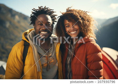 Happy African American couple in the mountains against the backdrop of a tent camp. Young international family is engaged in a mountain hike. Active lifestyle, tourism and vacation concept. Happy African American couple in the mountains against the backdrop of a tent camp. Young international family is engaged in a mountain hike. Active lifestyle, tourism and vacation concept. 107930796