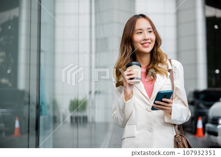 An Asian woman holds a paper cup of hot coffee while using her smartphone to stay connected with colleagues and clients. She is a savvy businesswoman. An Asian woman holds a paper cup of hot coffee while using her smartphone to stay connected with colleagues and clients. She is a savvy businesswoman. 107932351